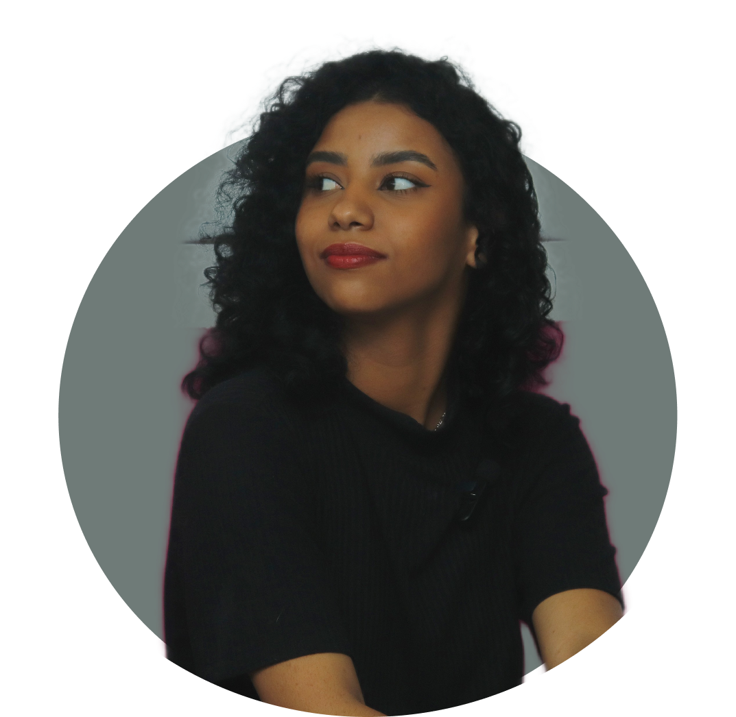 Jovem de cabelos cacheados pretos e batom vermelho, usando blusa preta, olha levemente para o lado com expressão confiante e serena. Ela está em frente a um fundo cinza escuro em formato circular. A jovem é Ellana Santos.